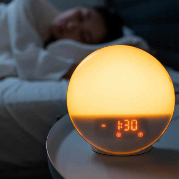 Réveil simulateur d'aube diffusant une lumière chaude orangée sur une table de nuit avec une personne qui dort en arrière-plan