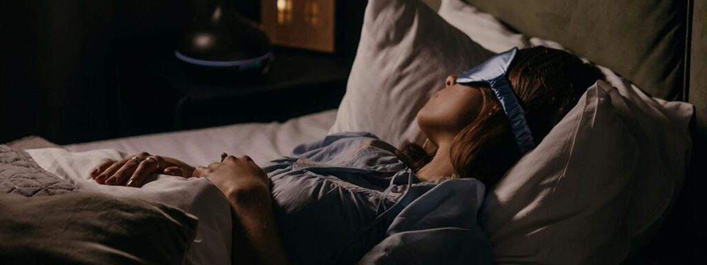 Femme endormie portant un masque de sommeil en soie bleue avec un diffuseur d'huiles essentielles en arrière-plan